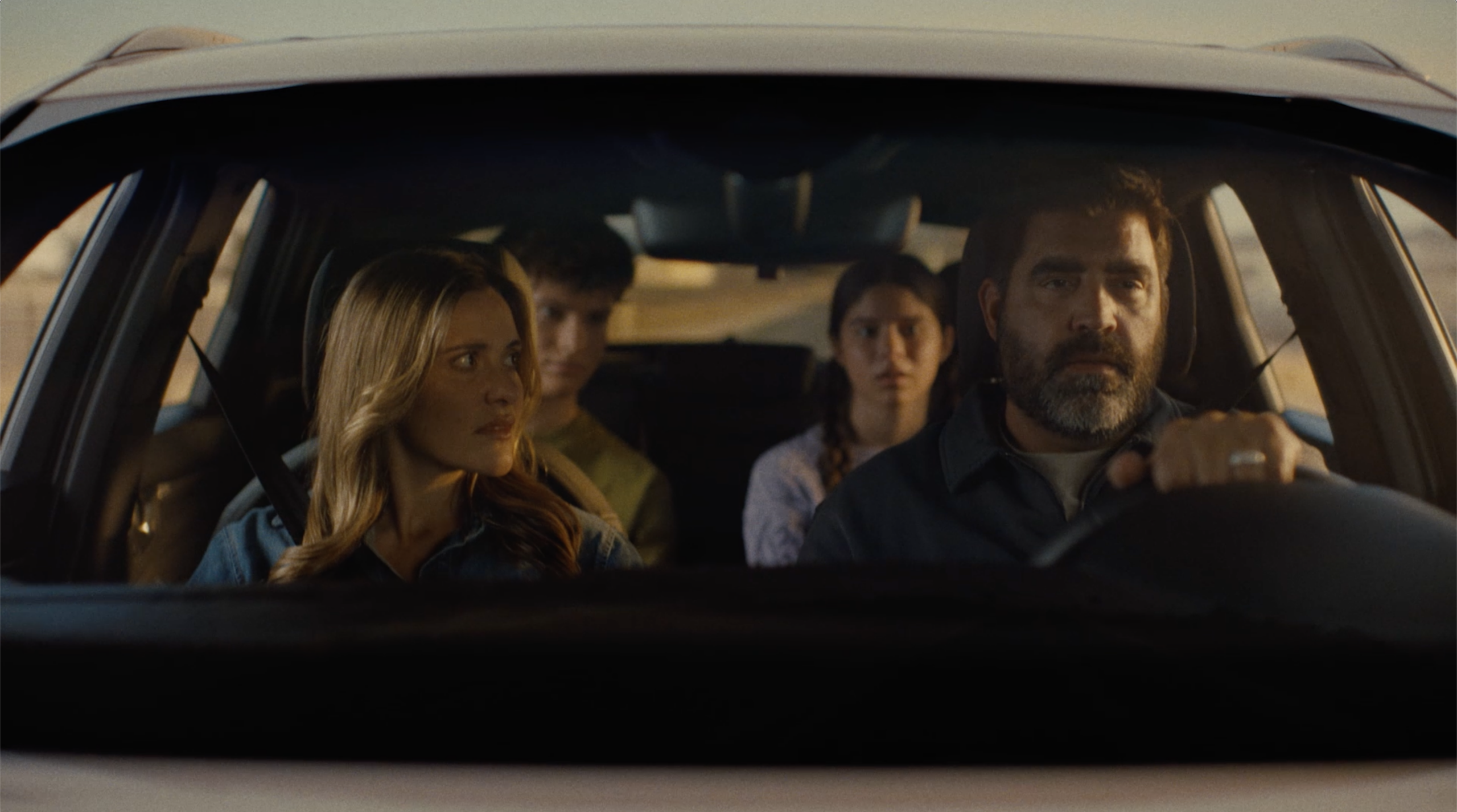 A family of four is tightly seated in a car. The father, driving, looks focused, while the mother appears concerned. The two teenagers in the back are serious. The scene is tense and somber.