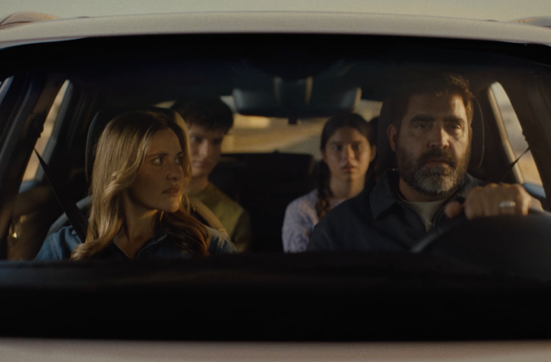 A family of four is tightly seated in a car. The father, driving, looks focused, while the mother appears concerned. The two teenagers in the back are serious. The scene is tense and somber.